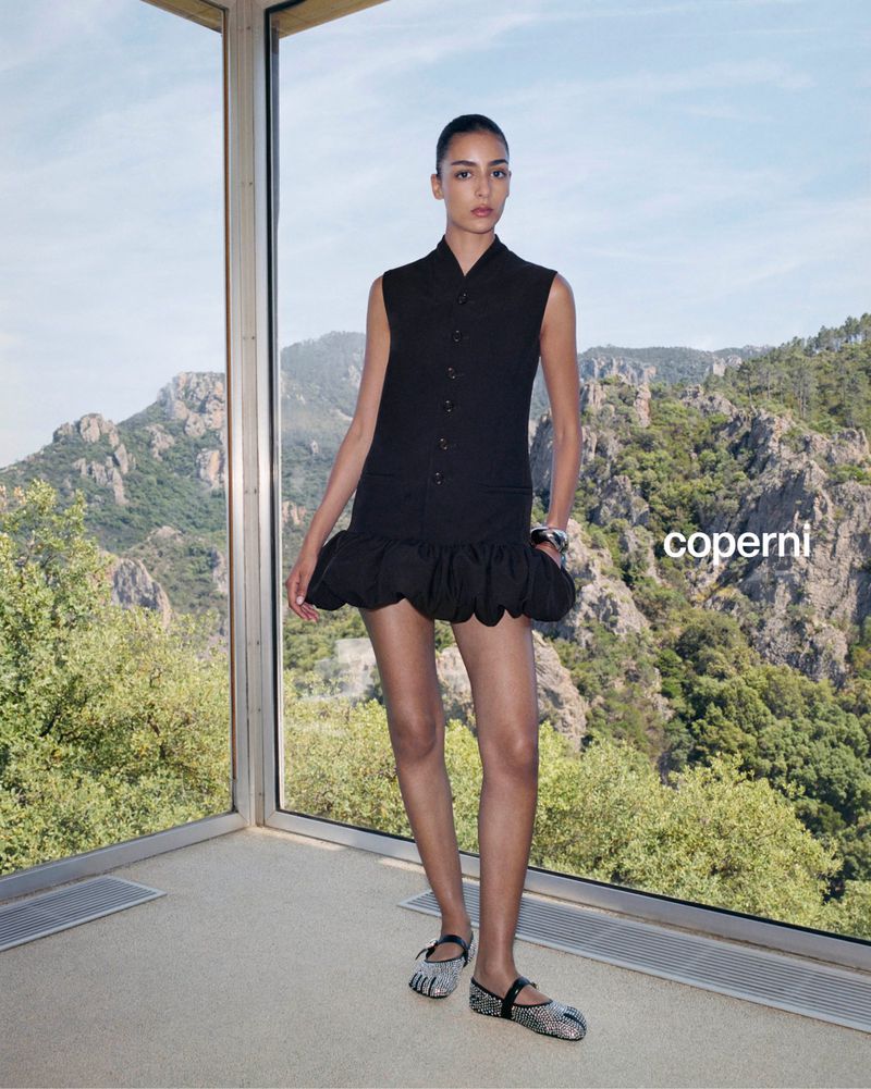 Indoor fashion shot of a woman posing in a black dress and shimmering flats next to a window with a mountain landscape view.