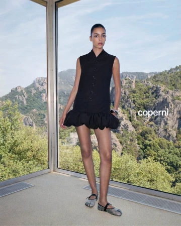 Indoor fashion shot of a woman posing in a black dress and shimmering flats next to a window with a mountain landscape view.