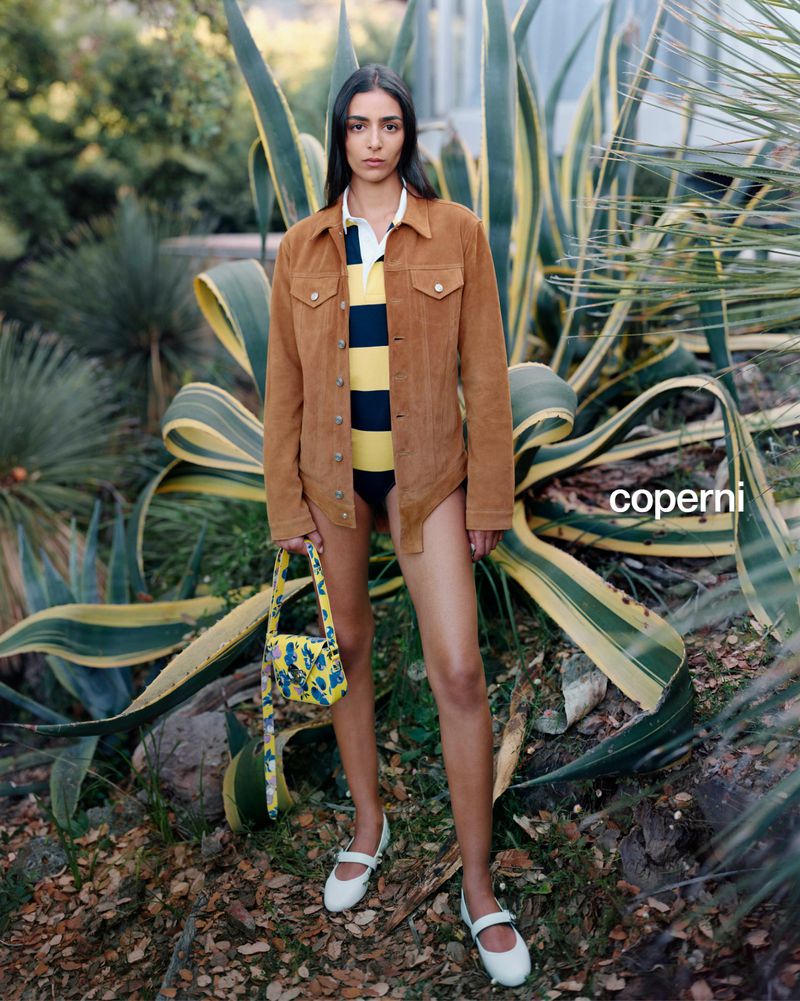 An adult woman poses in an outdoor setting, wearing a jacket, top, and white shoes, holding a handbag, with greenery in the background.