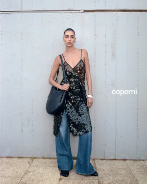 Model wearing a sequined slip dress over denim jeans with a black leather shoulder bag against a textured concrete wall.