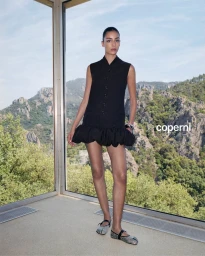 Model Nora Attal in a black sleeveless dress with a ruffled bubble hem standing before a scenic mountain window view.