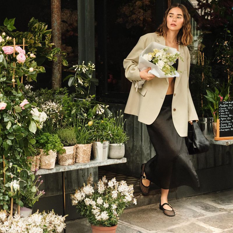 A stylish woman in a blazer holds a bouquet of flowers while walking outside a florist shop on a city street during the daytime.