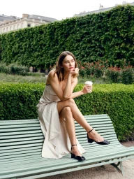 Model seated on a green park bench wearing a white dress and black heels, holding a coffee cup outdoors.