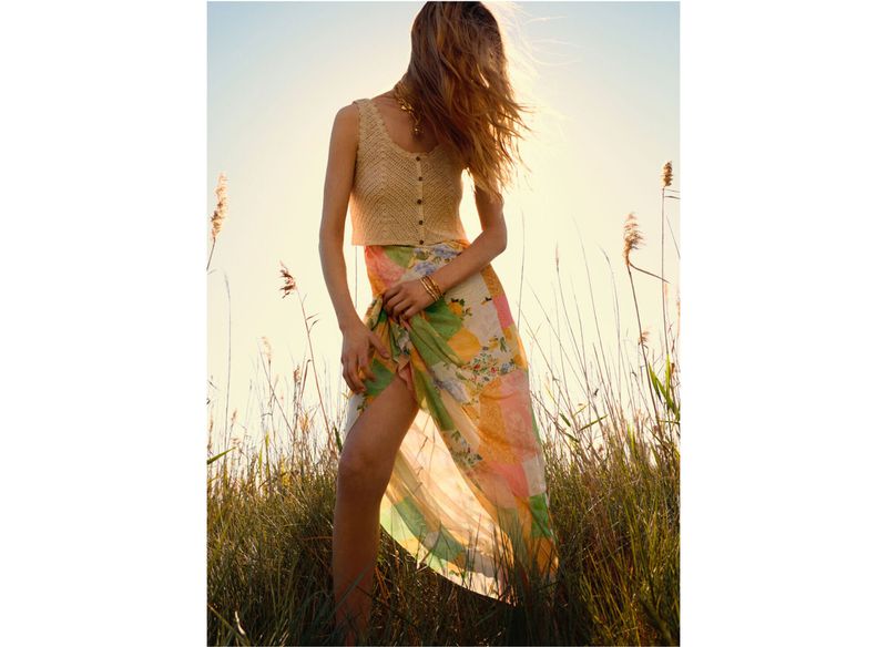 Woman in a knit top and colorful sarong stands in tall grassy field against the sun on a bright day, posing for a fashion shot.