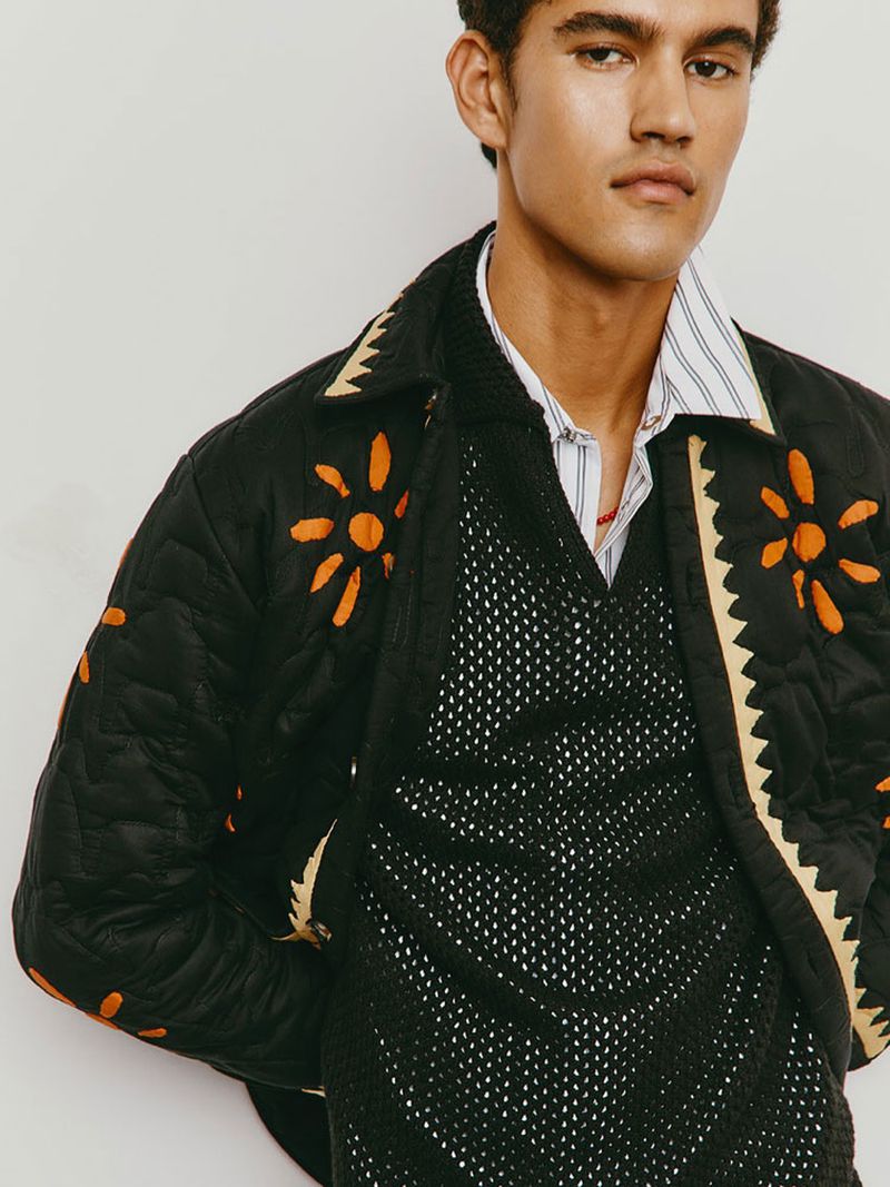 A stylish adult male poses indoors, wearing a black quilted jacket with orange floral embroidery, paired with a black mesh shirt and striped collared shirt.