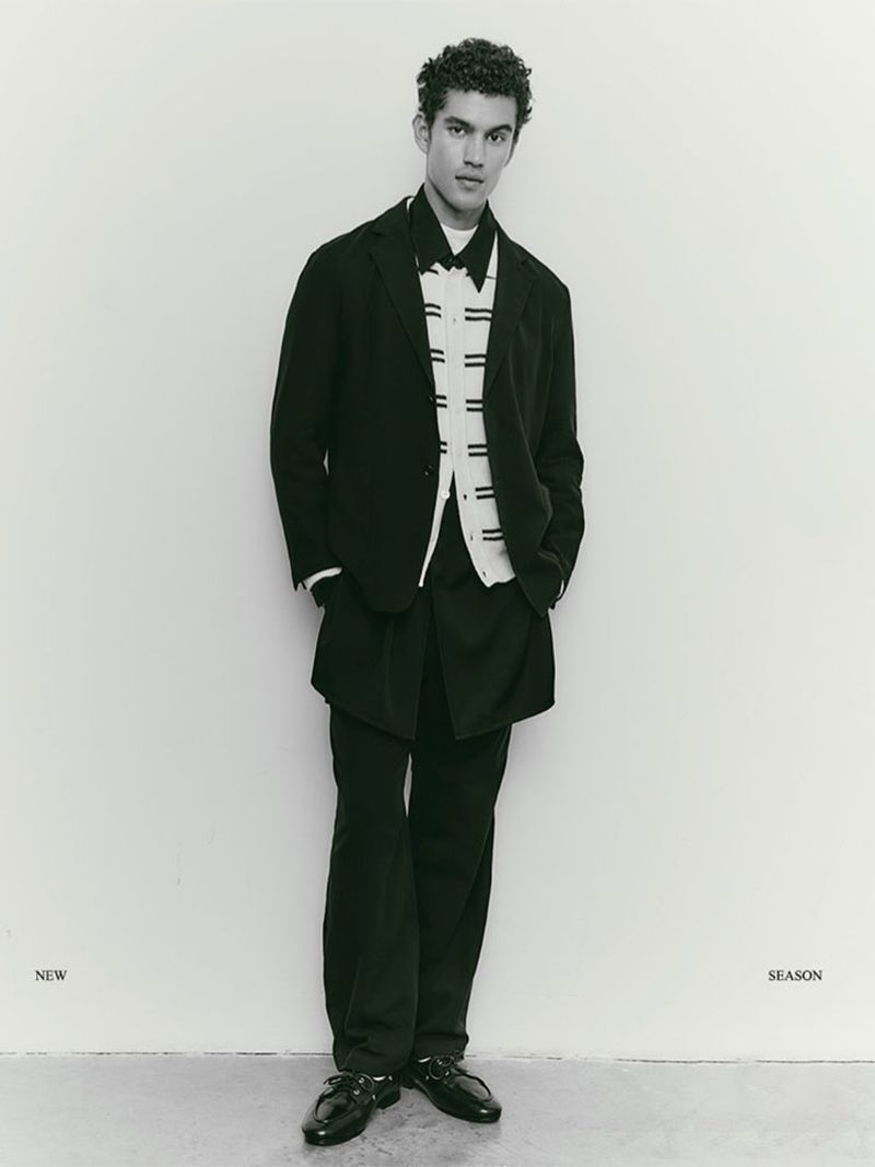 A stylish adult male stands indoors against a light wall, wearing a black blazer, matching slacks, bow tie, and patent leather shoes.