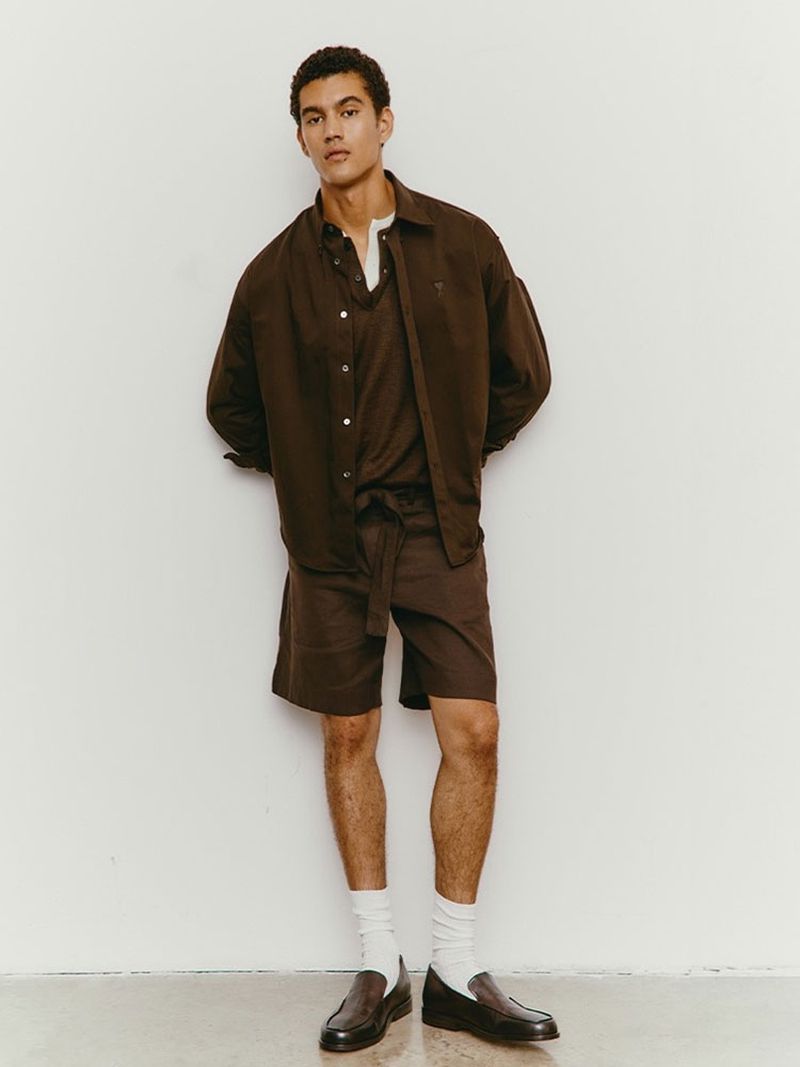 A relaxed studio portrait of a young adult male wearing a brown button-down shirt, matching shorts, white socks, and black loafers.