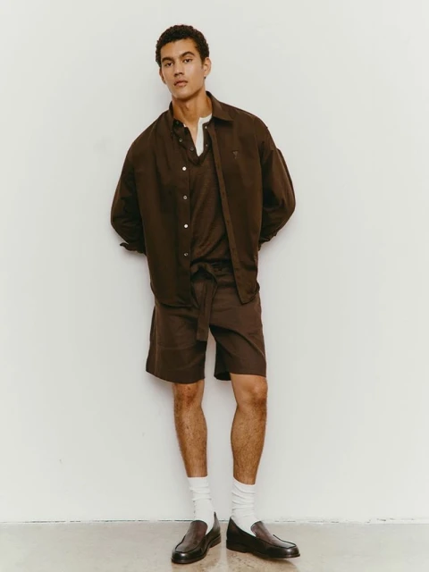 A relaxed studio portrait of a young adult male wearing a brown button-down shirt, matching shorts, white socks, and black loafers.