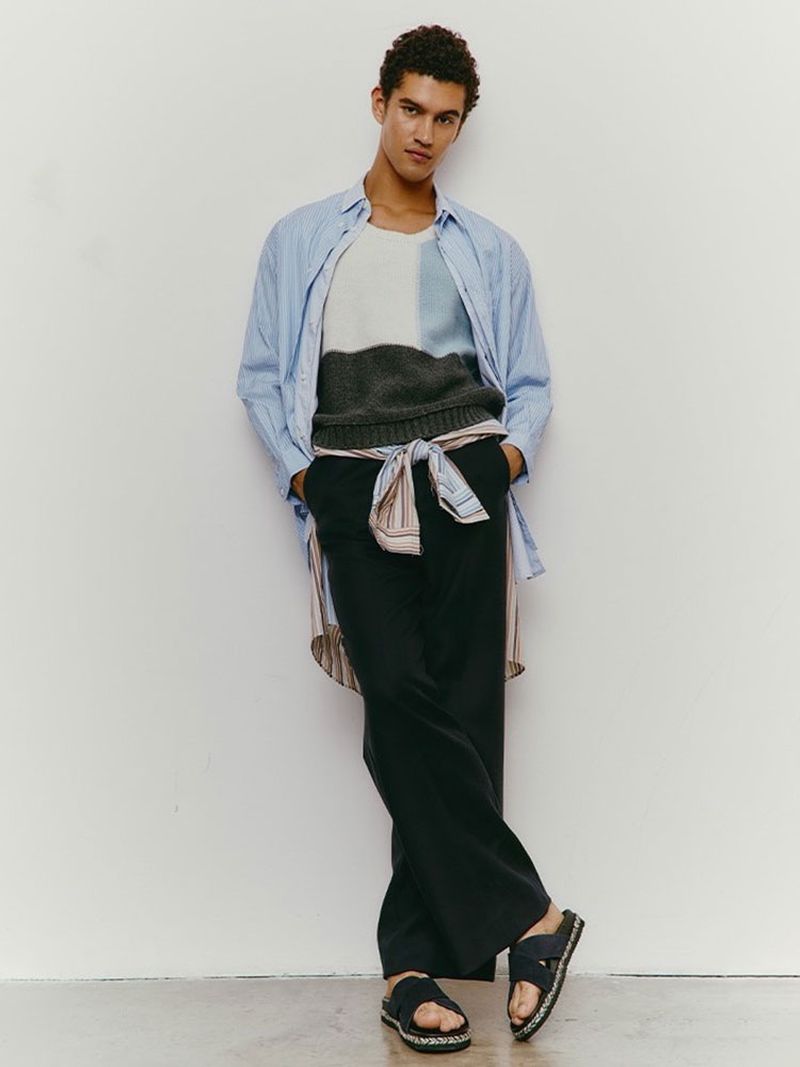 Male fashion model poses against a white wall, wearing a blue striped shirt, color-blocked sweater, black pants, and black sandals in a studio.
