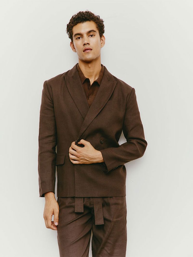 An adult male model wearing a double-breasted brown linen suit, posing against a plain white background in a fashion studio shoot.