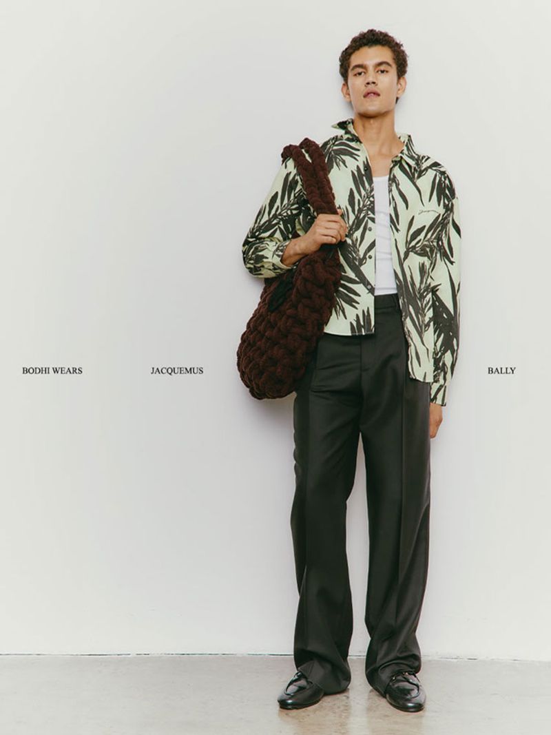 Male fashion model in a patterned shirt, trousers, and loafers, carrying a chunky woven bag, posing against a white wall.