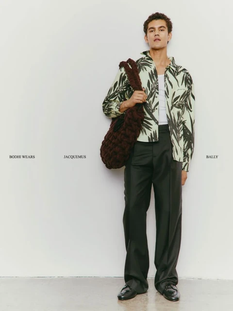 Male fashion model in a patterned shirt, trousers, and loafers, carrying a chunky woven bag, posing against a white wall.