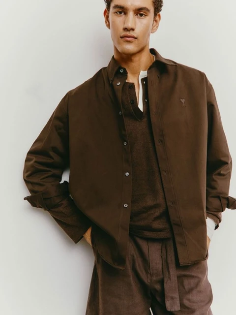 Young man in a brown collared shirt and brown linen pants poses in a studio, looking at the camera in a fashion shot.