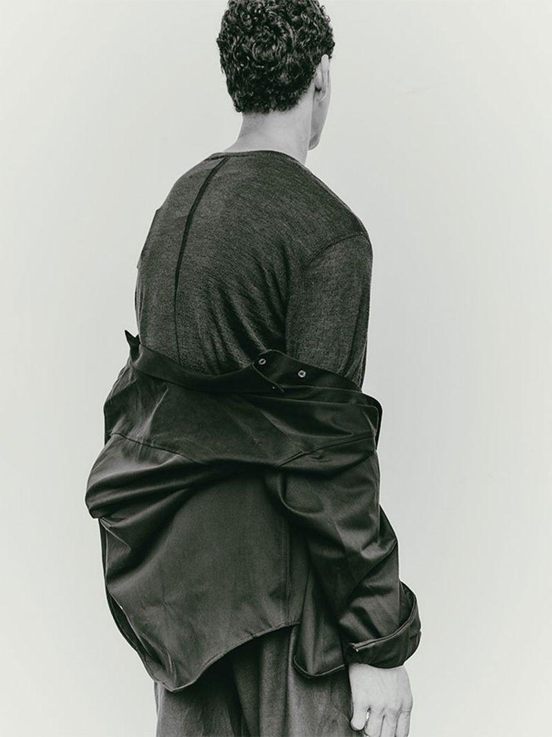 An adult male, seen from the back, wears a gray shirt and dark jacket against a plain white background in a studio fashion shot.