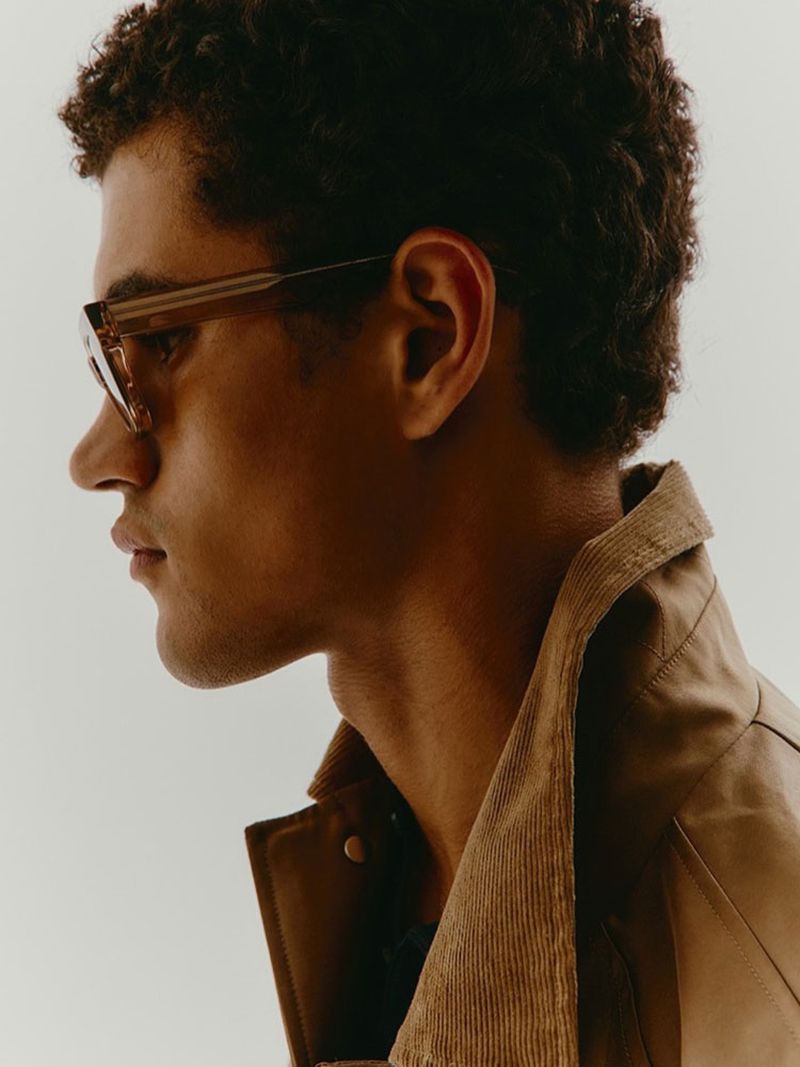 Portrait of a curly-haired man wearing sunglasses and a brown corduroy jacket against a white background in a studio setting.