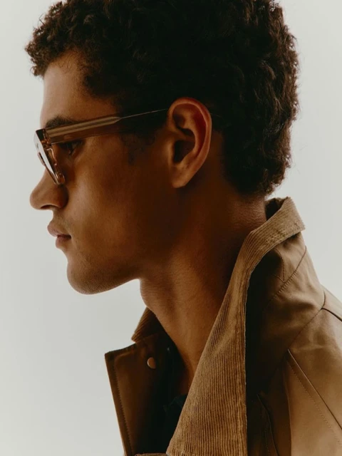Portrait of a curly-haired man wearing sunglasses and a brown corduroy jacket against a white background in a studio setting.
