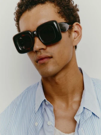 Close-up of a model wearing oversized square sunglasses and a striped shirt, posing against a neutral backdrop in a studio setting.
