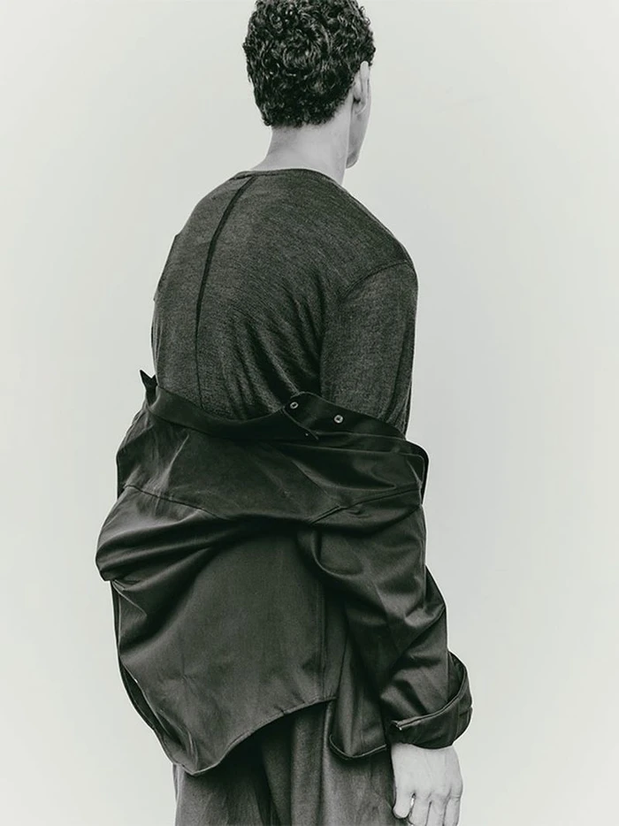 Model with dark curly hair seen from behind wearing a dark grey shirt and a black jacket draped over shoulders.