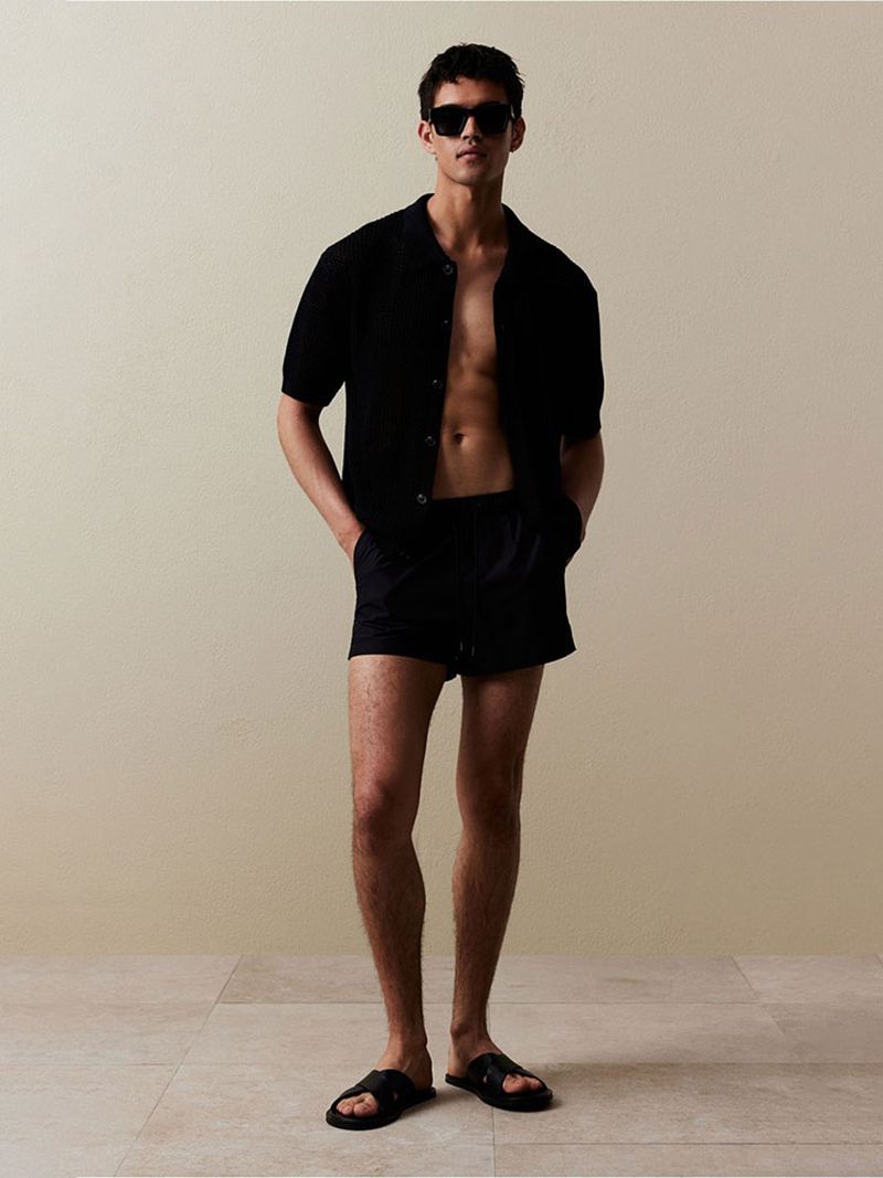 An adult male stands against a cream-colored background wearing a black button-down shirt, black shorts, sunglasses, and black sandals.