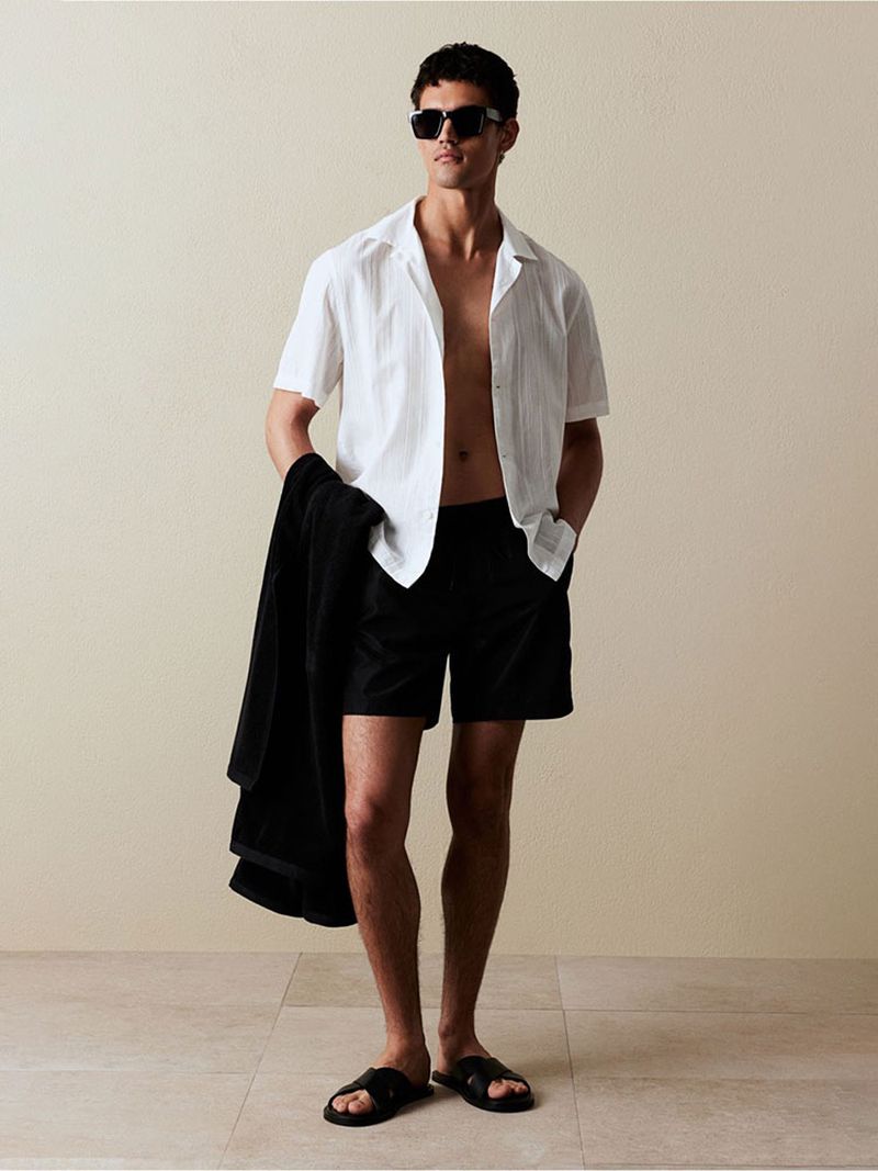 Man in casual white shirt and black shorts poses in front of neutral wall holding a black towel, wearing sunglasses and sandals.