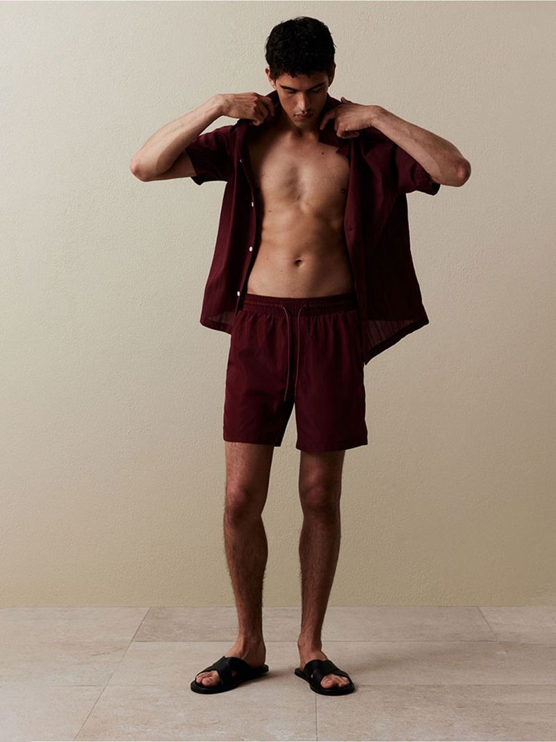 A fair-skinned male in burgundy shirt and shorts poses in a photo studio with a neutral background and wears black slide sandals.