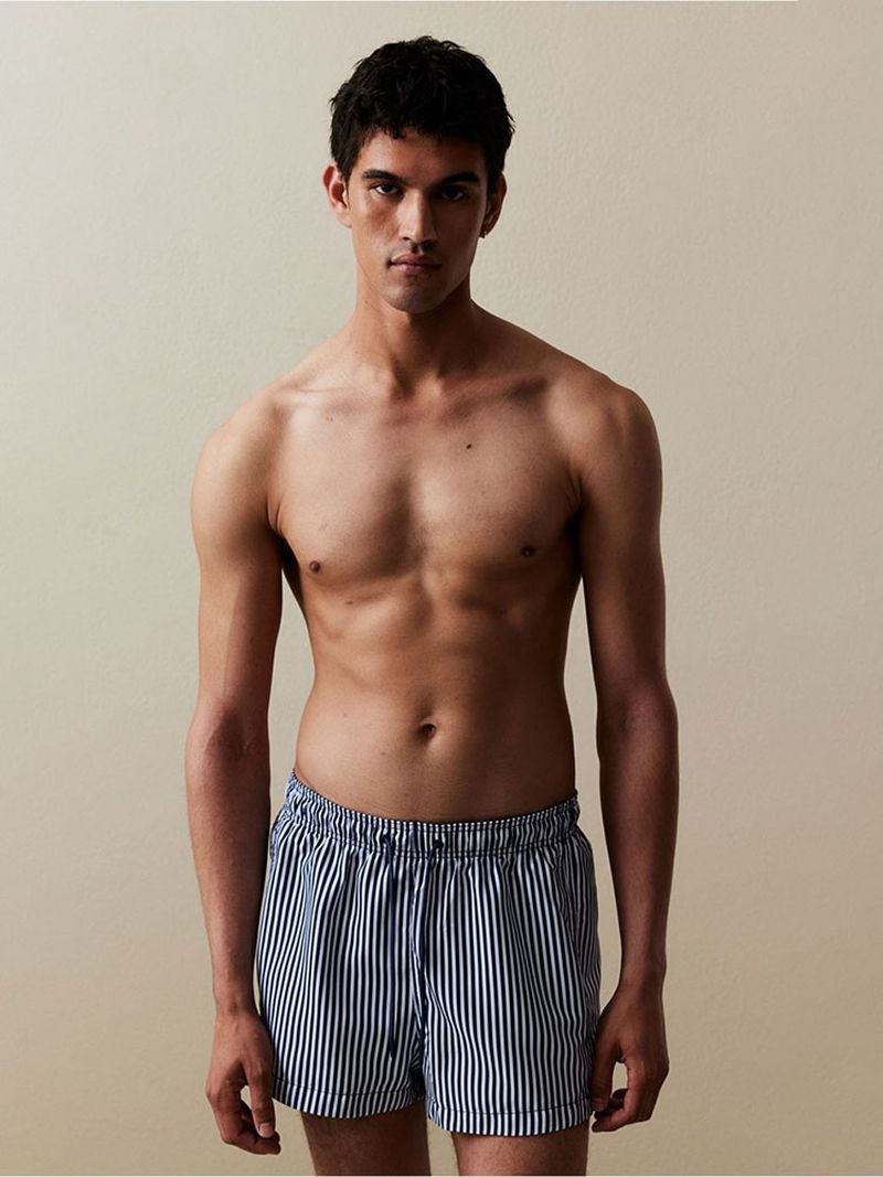 Adult male poses shirtless wearing striped boxer shorts in a studio setting with neutral-toned background, medium shot.
