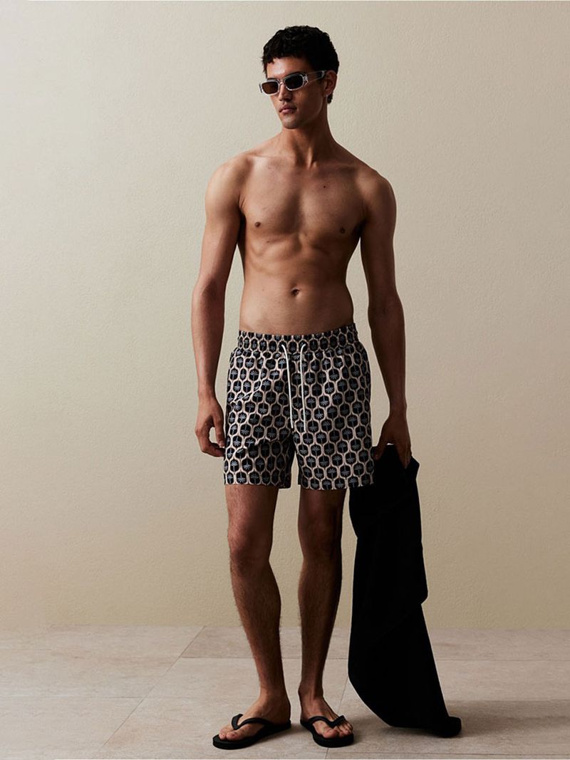 A man poses in printed swim trunks with a black jacket, sunglasses, and flip-flops against a neutral studio backdrop, fashion photography.