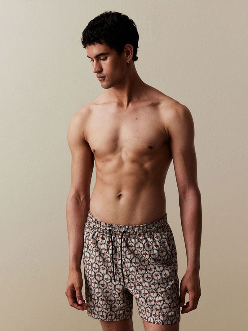 A shirtless man poses indoors wearing patterned swim trunks, gazing down with neutral expression. The shot is medium and studio-lit.