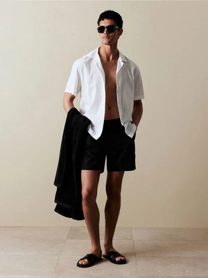 Male model wearing dark sunglasses, unbuttoned white shirt, and black swim shorts holding a dark towel against a neutral studio background