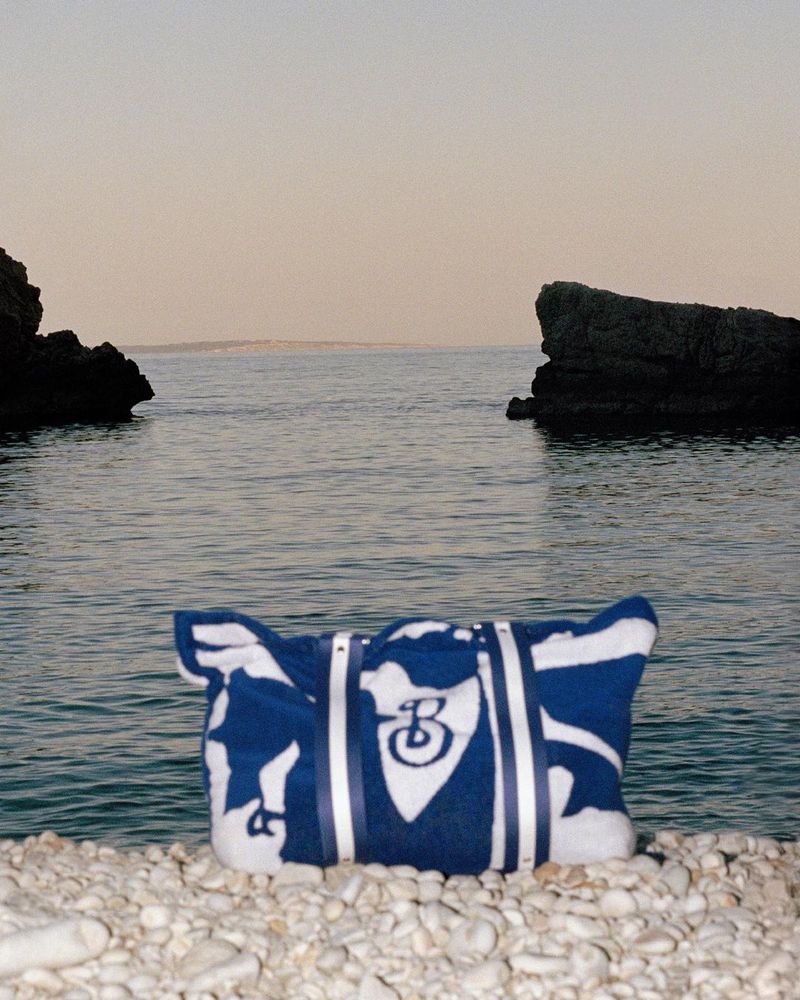 Blue and white beach bag on a pebbled shore with a backdrop of ocean and rocky cliffs under a serene sky at daytime.
