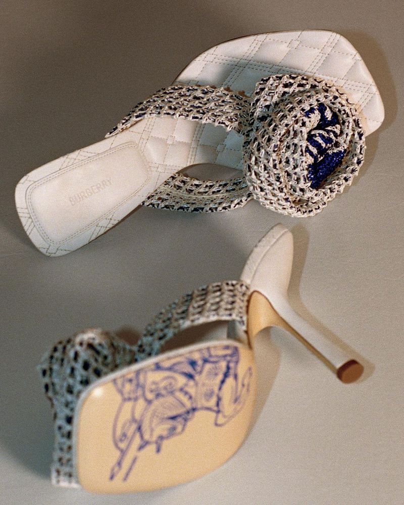 Elevated view of white high-heeled sandals with mesh straps, decorative rosette, and a blue design visible on the sole.