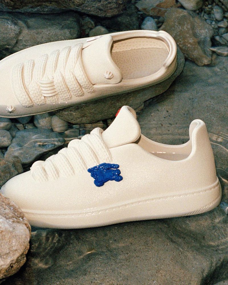 Two identical white sneakers with laces and blue logo on a rock and in shallow water in an outdoor setting, fashion still life. 