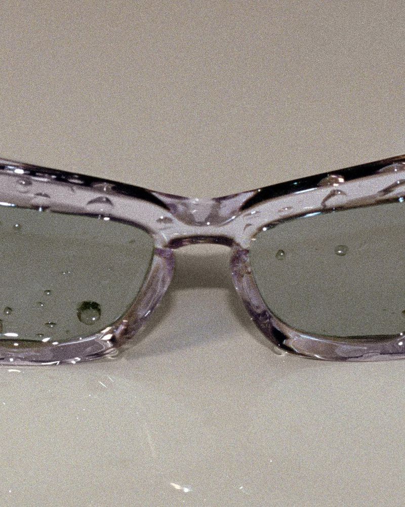 Close-up of transparent lilac sunglasses with water droplets on the lenses and frame, resting on a light-colored surface.