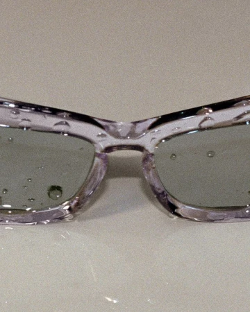Close-up of transparent lilac sunglasses with water droplets on the lenses and frame, resting on a light-colored surface.