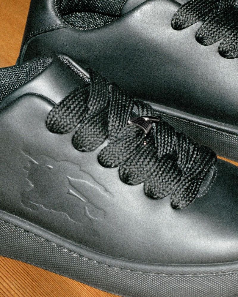 Close-up shot of sleek black leather sneakers with detailed laces and subtle embossed logo. The sneakers sit on a neutral surface.