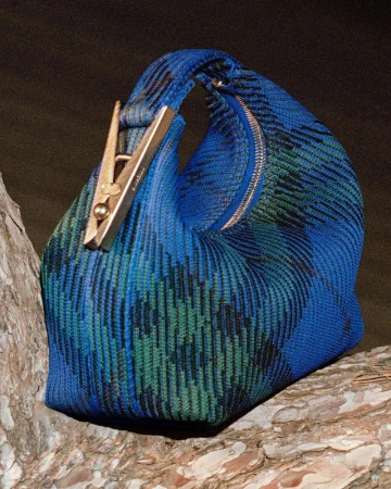 Close-up of a blue and green woven handbag on a textured surface, featuring a unique gold-colored clasp design element.