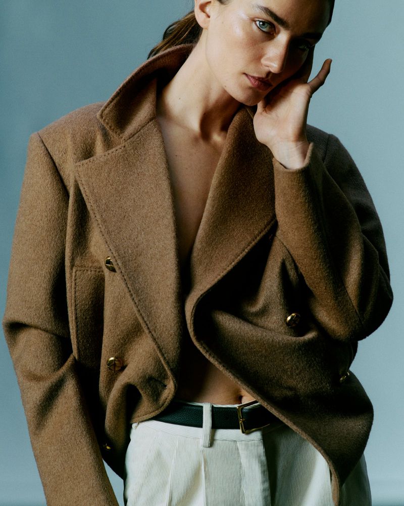 A woman with fair skin poses in a brown wool jacket with gold buttons and white pants against a light blue studio background.