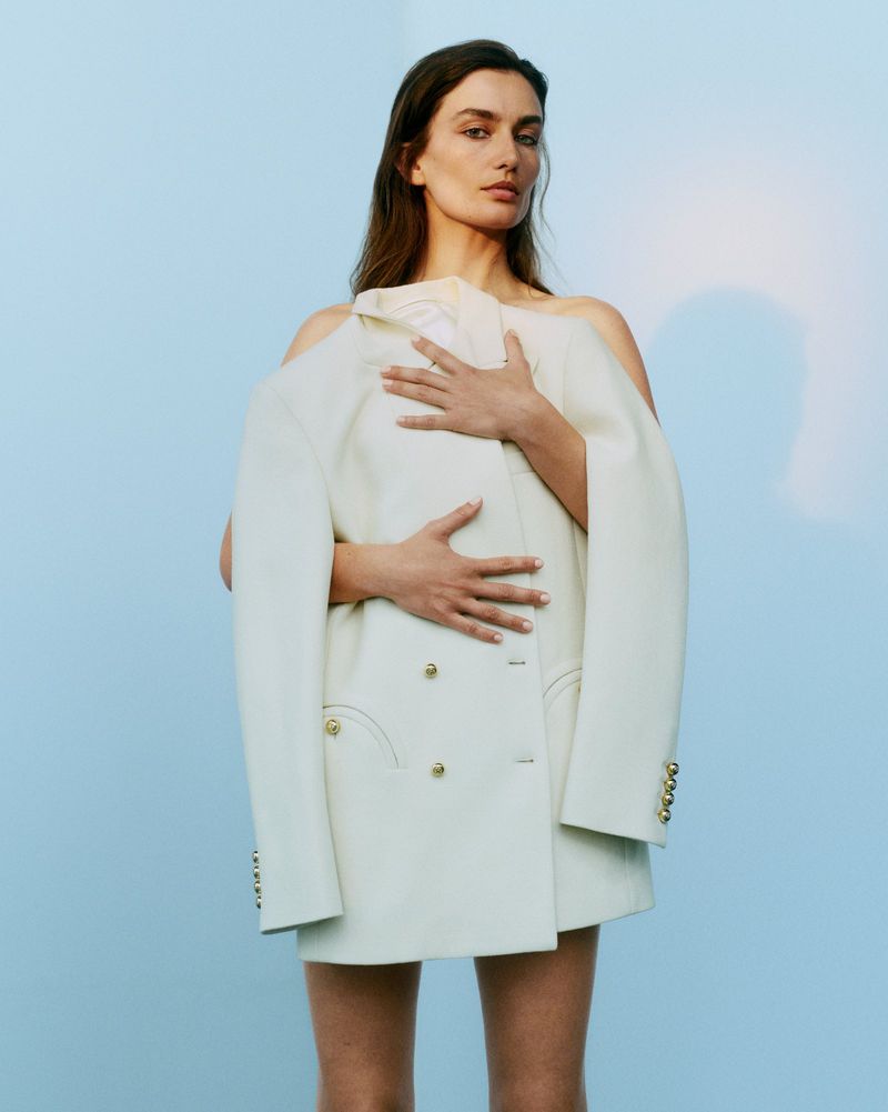A woman poses in front of a light blue background, holding a cream colored, oversized blazer across her chest, looking directly at the camera.