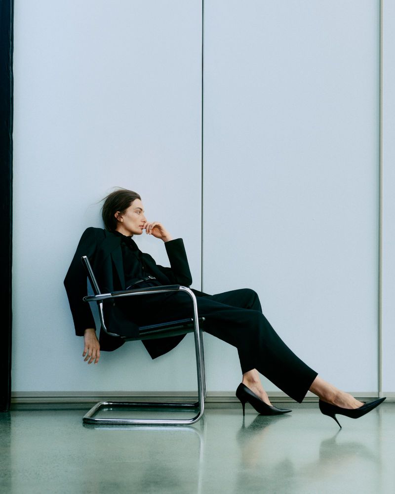 Elegant woman in black suit and heels sitting in a chrome chair against a white wall in a fashion-forward pose and setting.