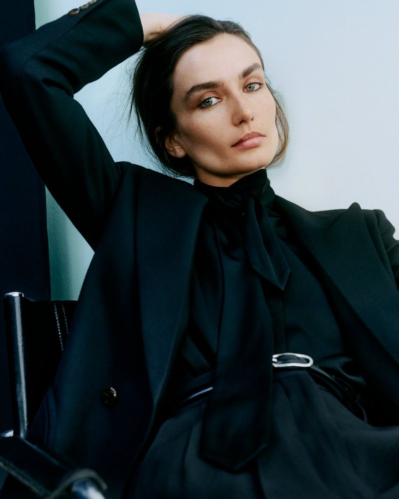Fashion portrait of a woman in a black pantsuit and pussy-bow blouse sitting on a black chair in front of a gray and black wall.