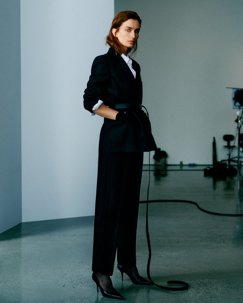 A female fashion model in a black blazer, white shirt, and black slacks strikes a full-length pose in a studio with lighting equipment.