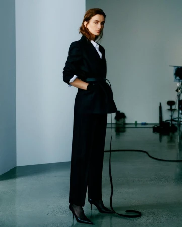 A female fashion model in a black blazer, white shirt, and black slacks strikes a full-length pose in a studio with lighting equipment.