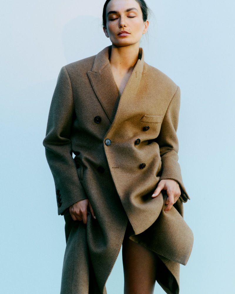 Woman in a tan, oversized double-breasted blazer posing against a light blue background in a fashion-forward portrait.