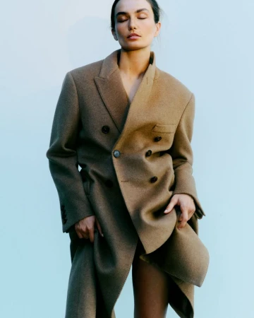 Woman in a tan, oversized double-breasted blazer posing against a light blue background in a fashion-forward portrait.