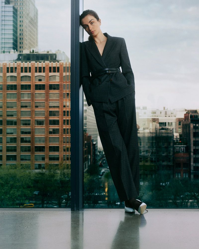 Woman in a dark grey suit and sneakers leans against a window in an urban environment, skyscrapers visible in the background. Fashion shot.
