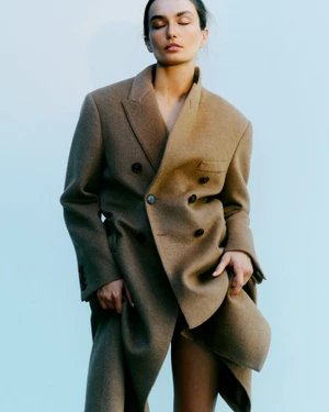 Model Andreea Diaconu wearing a structured camel double-breasted coat against a pale blue seamless backdrop.