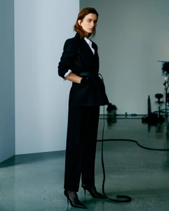 Model Andreea Diaconu in a black blazer and trousers holding hands in pockets against a minimalist white studio background