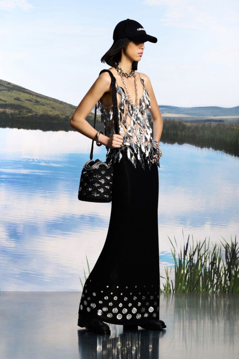 Fashion model poses outdoors wearing a metallic sequin top, long black skirt, hat, and bucket bag against a lake background.