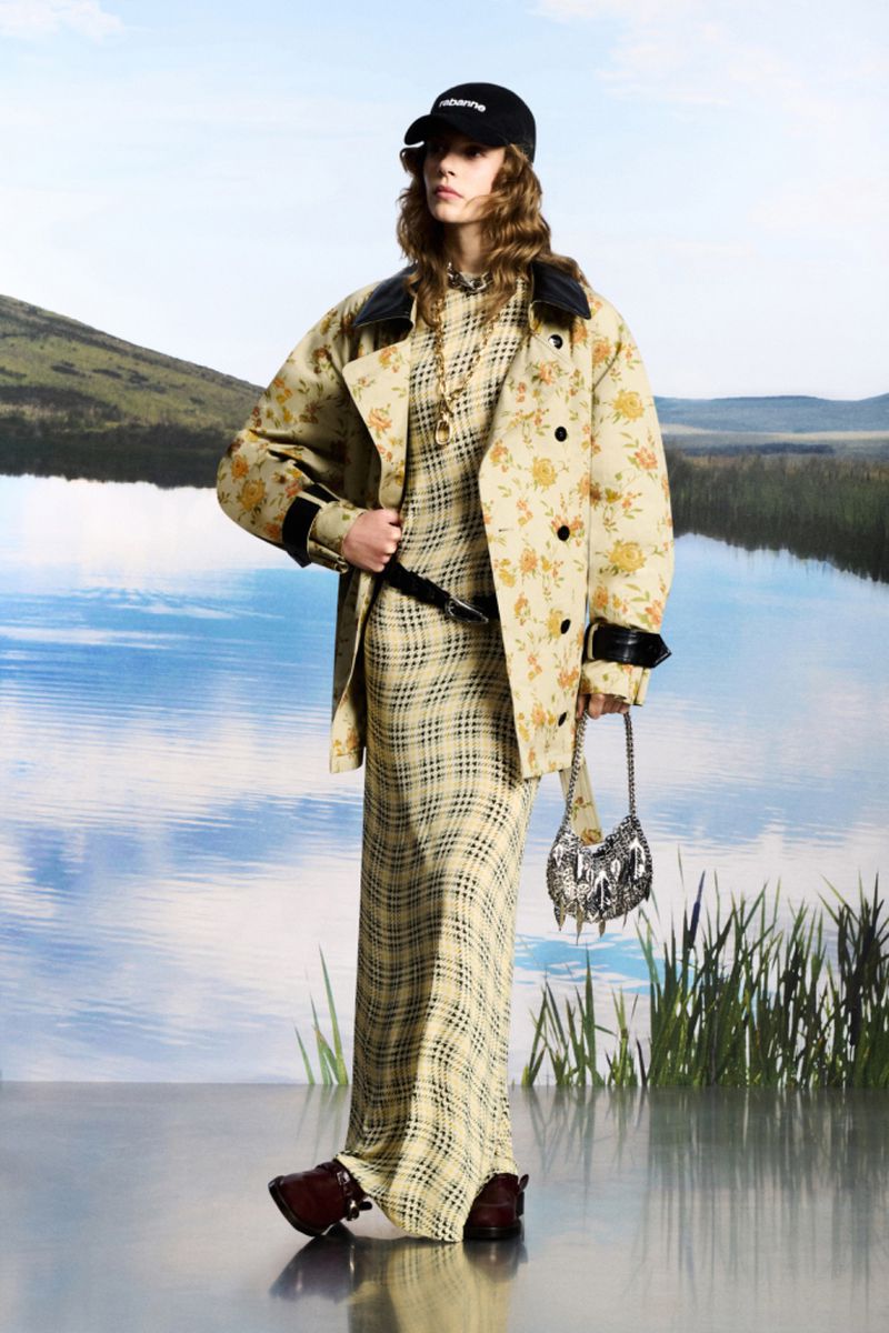 Adult female fashion model poses in a floral trench coat, plaid dress, cap, handbag, and boots against a blurred nature background.