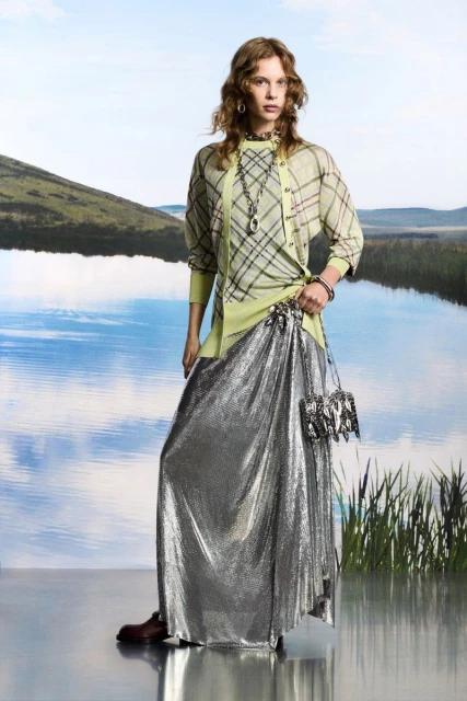 Fashion photo of a woman posing in front of a lakeside landscape, wearing a green plaid cardigan and silver metallic skirt, studio setting.